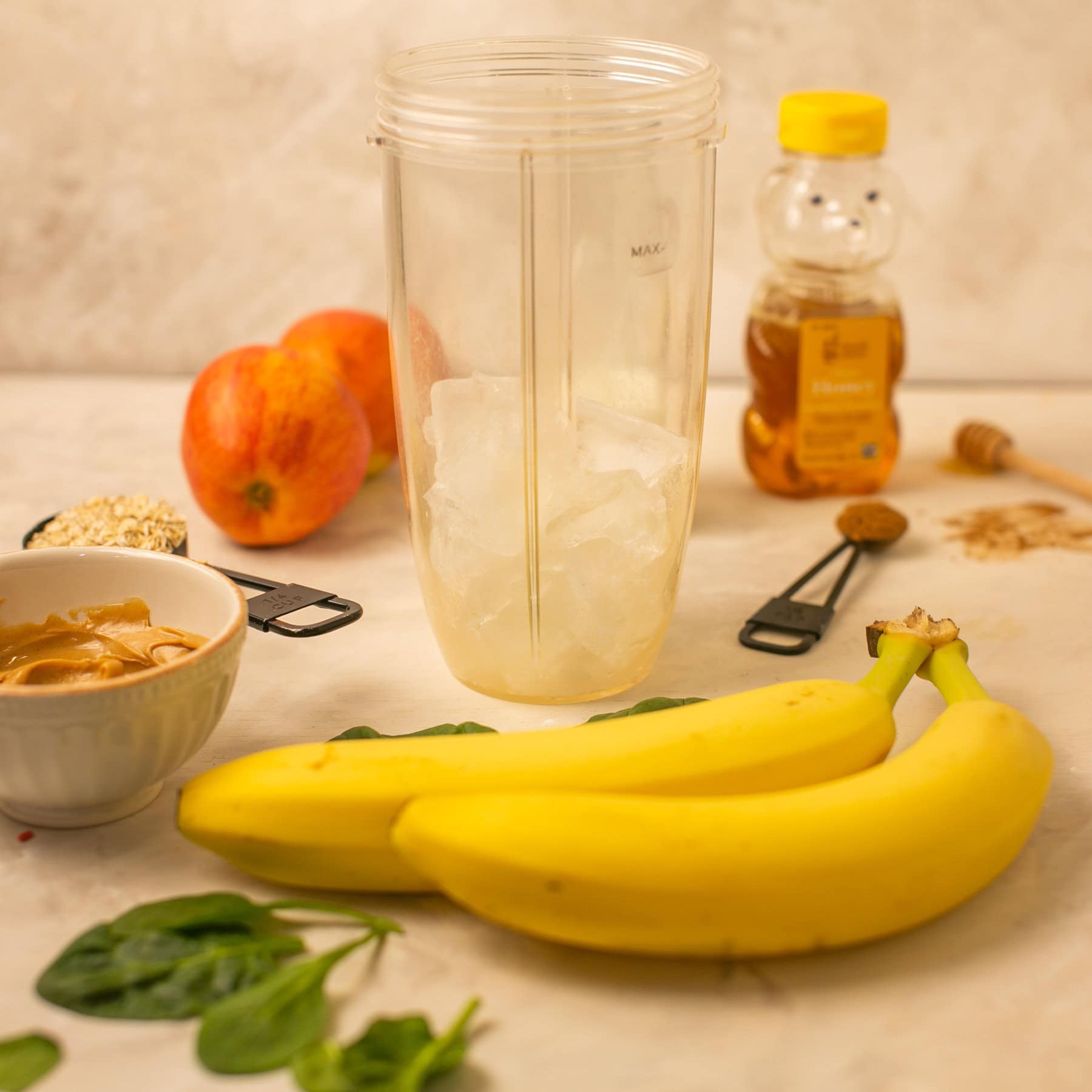A picture showing the beginning step of the process with a cup of ice, peanut butter, bananas, spinach, cinnamon, and honey. 
