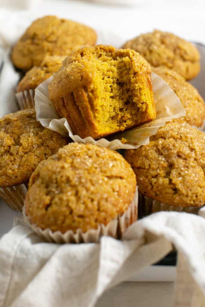 A bunch of pumpkin spice muffins sitting in a pan. One muffin is on top with a bite taken out. 