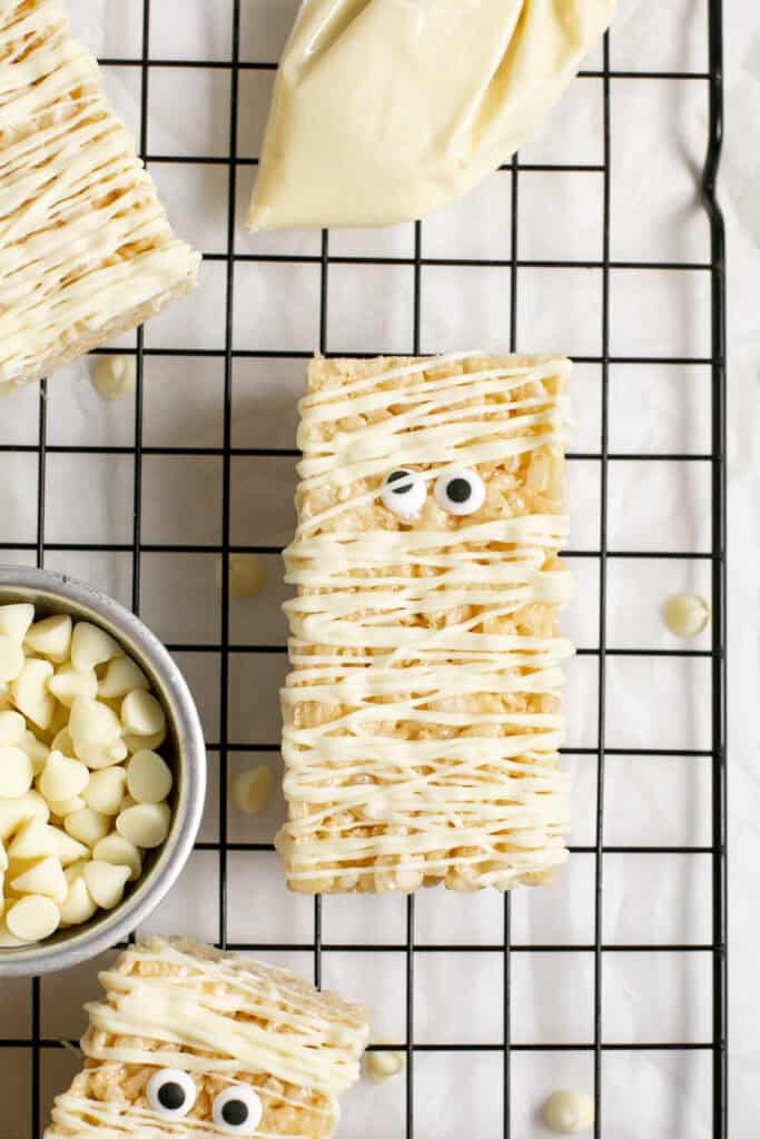 the mummy marshmallow treats sitting on a cooling rack with some white chocolate chips. 